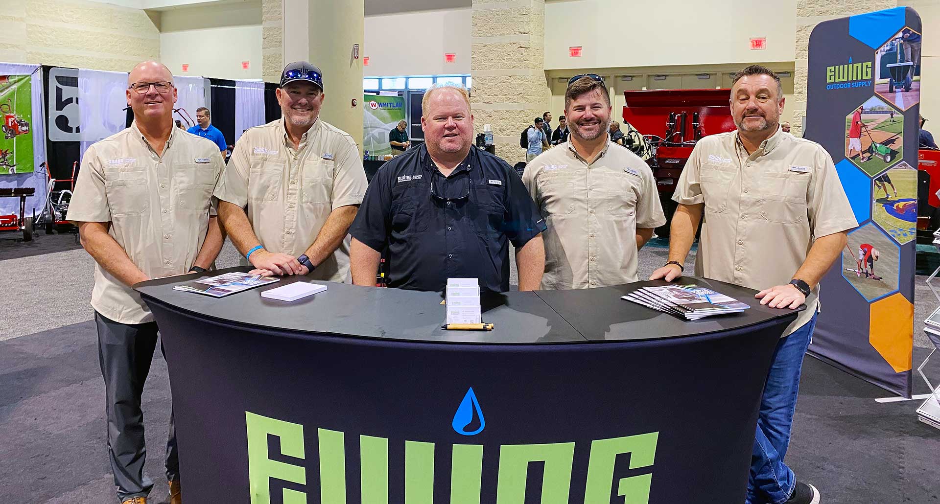 Five men in Ewing black and tan Ewing shirts behind a black counter with the Ewing logo on it and a sign behind them with a Ewing logo and images of contractors working.