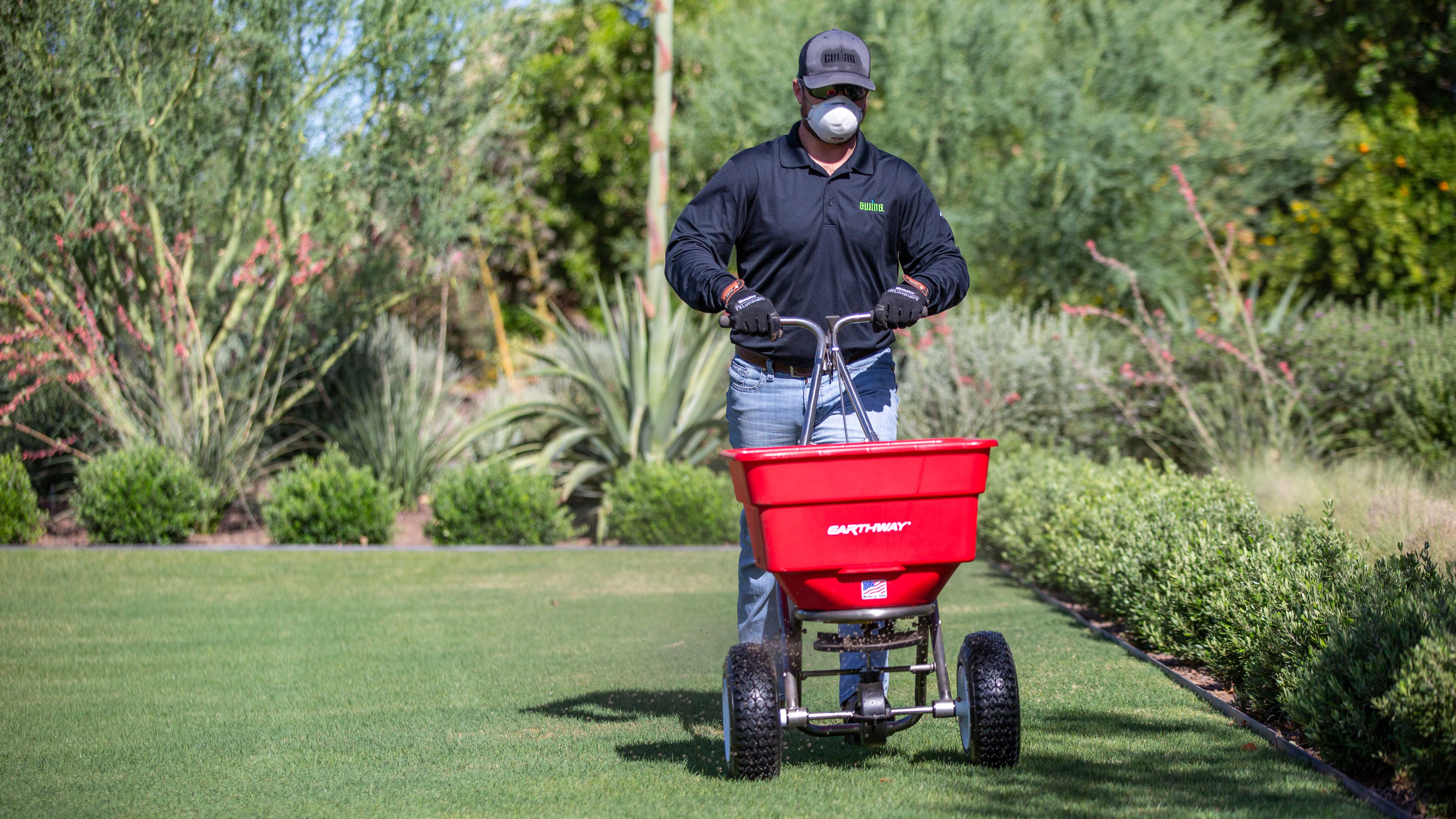 Contractor wearing a gray hat and long-sleeved black shirt pushing a red EarthWay broadcast spreader on a green lawn.