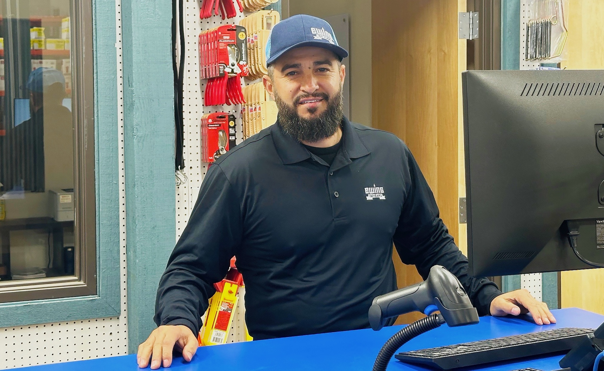 Ewing Windsor Branch Manager Nile Barajas at the blue counter, wearing a black shirt and blue hat, with products on the wall behind him.