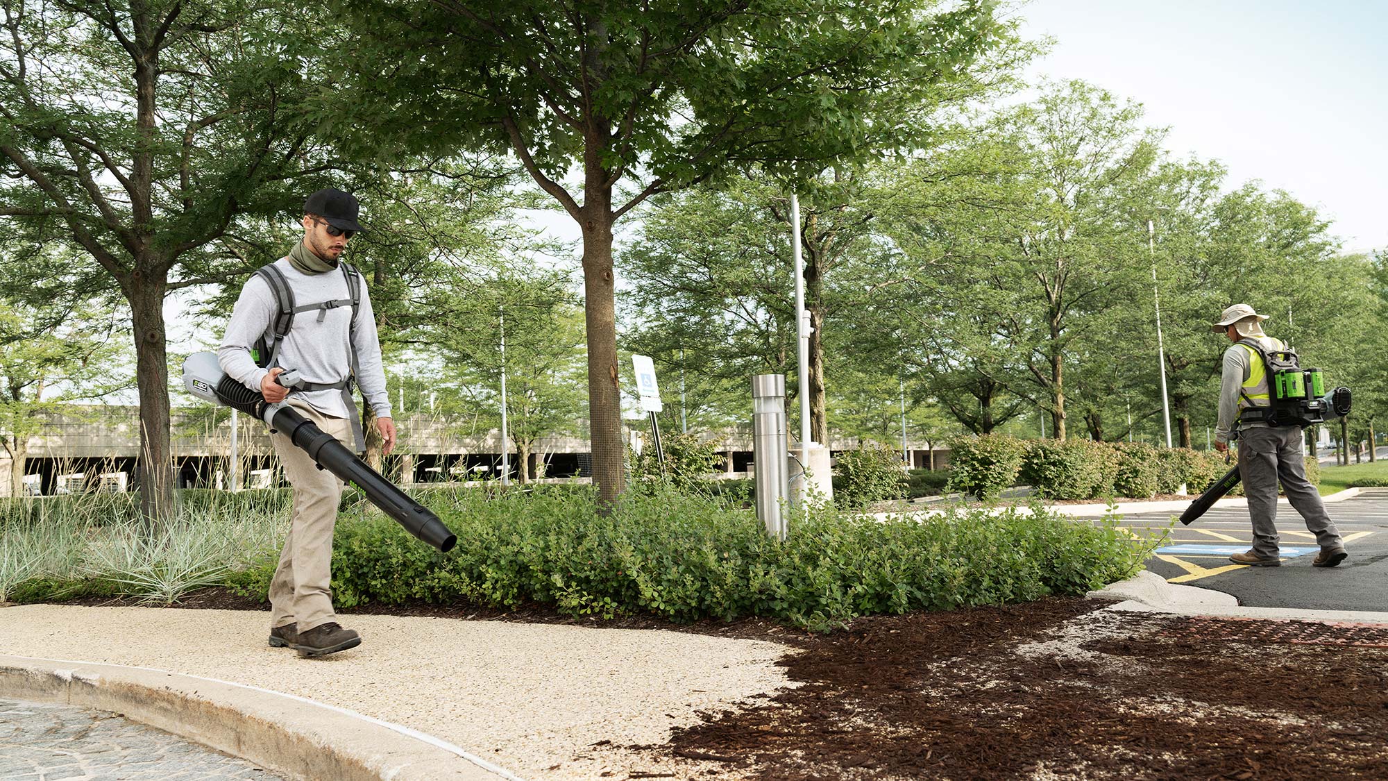 Two landscape contractors use battery-powered backpack blowers to clear off a sidewalk and parking area.