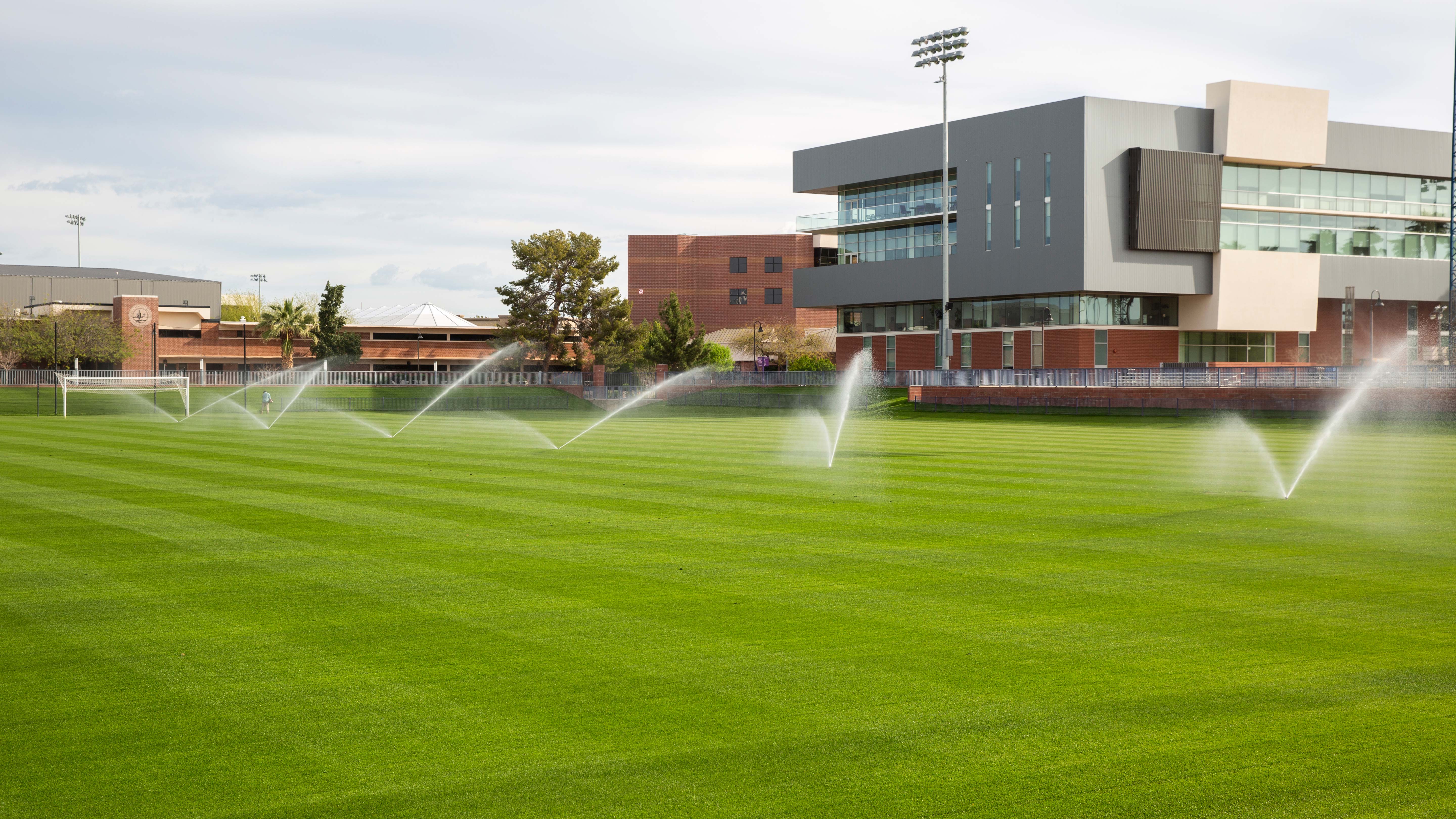 Green manicured sports field with sprinklers in front of modern gray and rust-colored buildings.