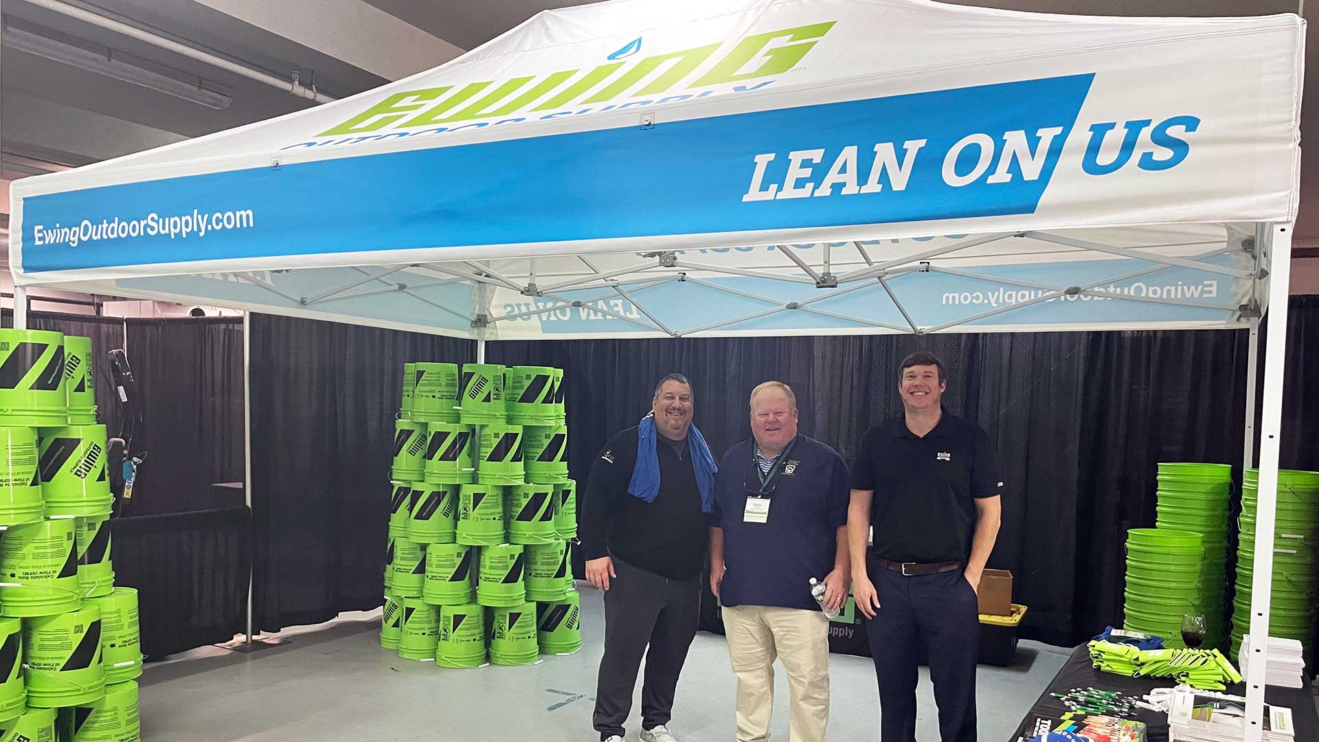 Three men in black shirts standing under Ewing Outdoor Supply canopy with green buckets and items on a table.